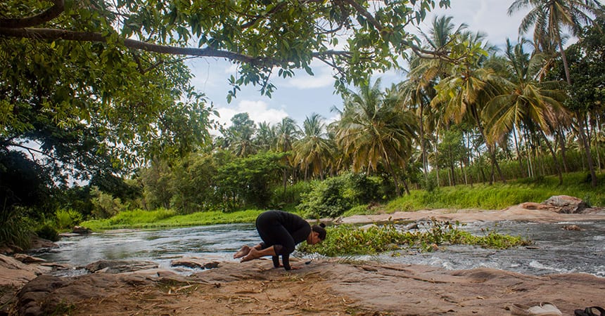 crow pose (bakasana) variation performed outdoors on a rock by a rushing river surrounded by trees in Karnataka